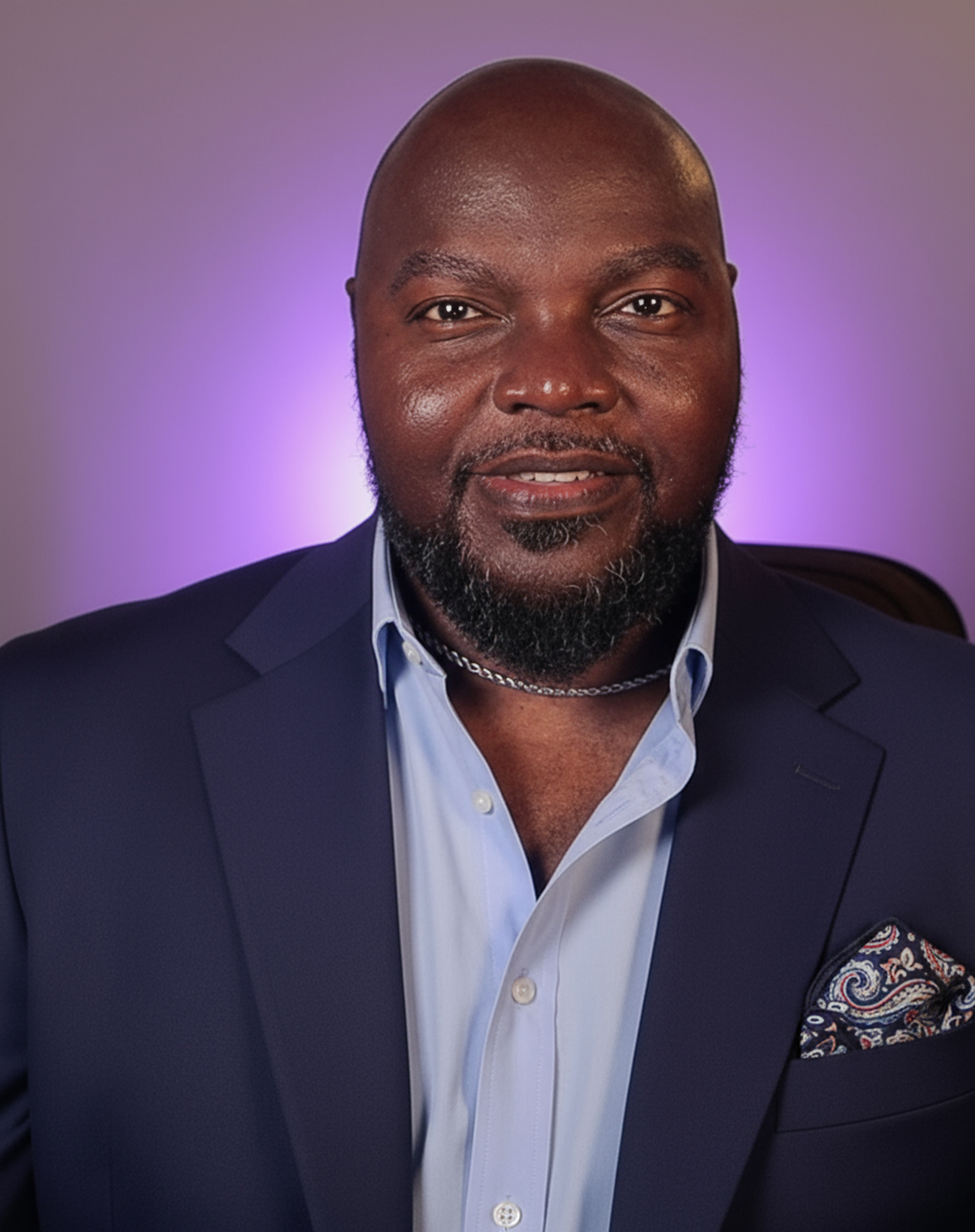 LaMondré Pough, a bald Black man with a neatly groomed beard, wearing a navy suit jacket, light blue shirt, and paisley pocket square, smiles confidently against a softly lit purple background.