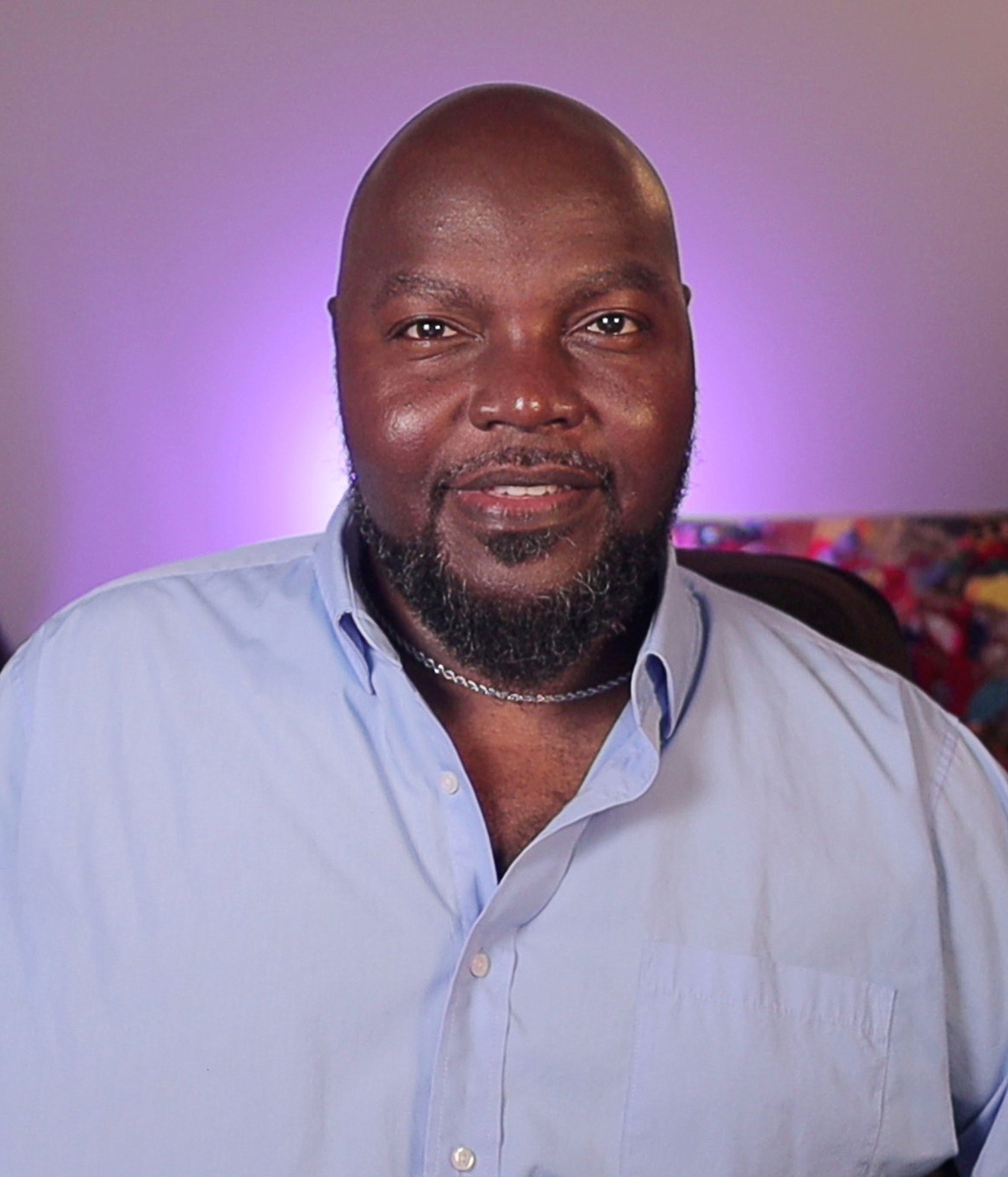 LaMondré Pough smiling in a light blue button-down shirt with a soft purple background glow, presenting a warm and approachable headshot.