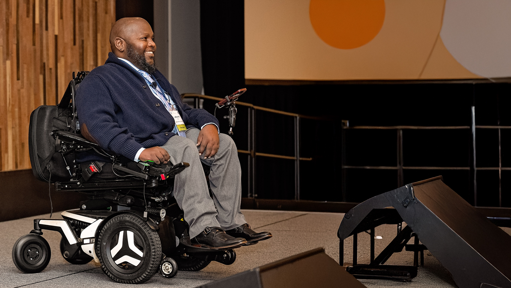 LaMondré Pough smiling while seated in a power wheelchair on stage, wearing a navy sweater and gray pants, speaking at a conference with a wooden panel wall and presentation screen in the background.” Five